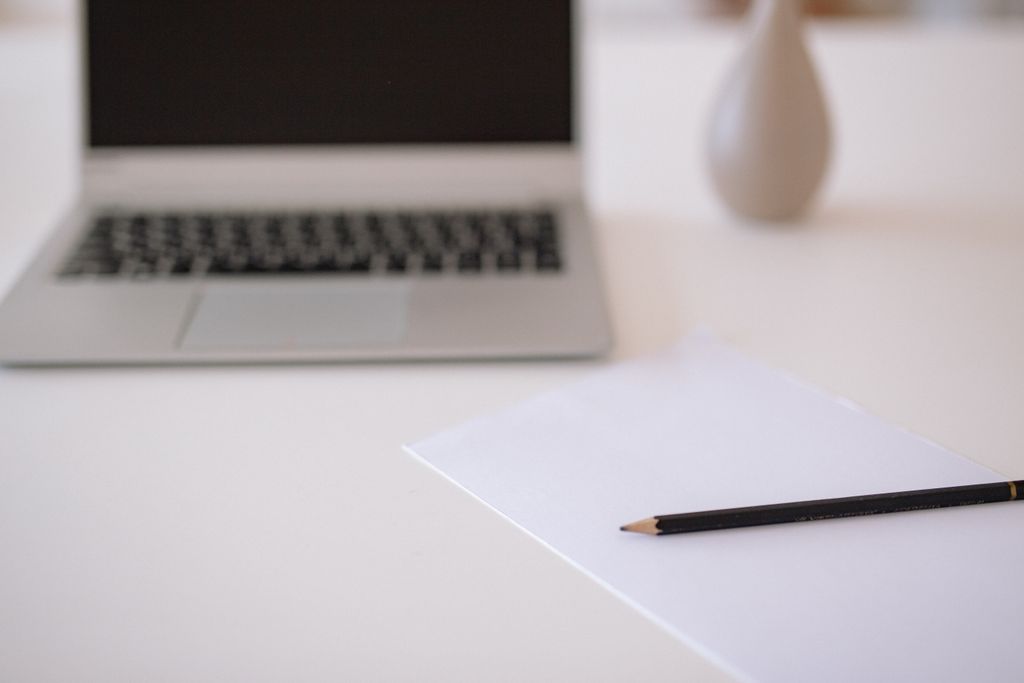 A minimalistic workspace featuring a laptop, blank paper, and pencil on a white table.